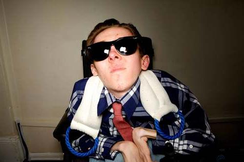 A young male with light skin using a wheelchair and wearing a blue shirt, a red tie, and sunglasses. Behind him, there is a tan wall. 